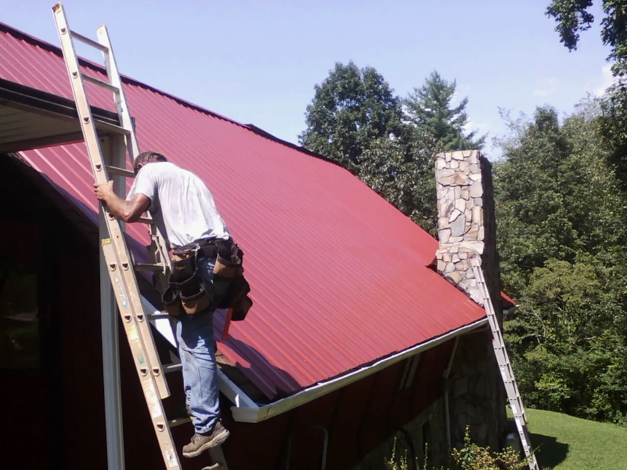 Roofer on a ladder beside a red metal roof and stone chimney — Soffit & Fascia Repair by Roofing Brothers in Altoona